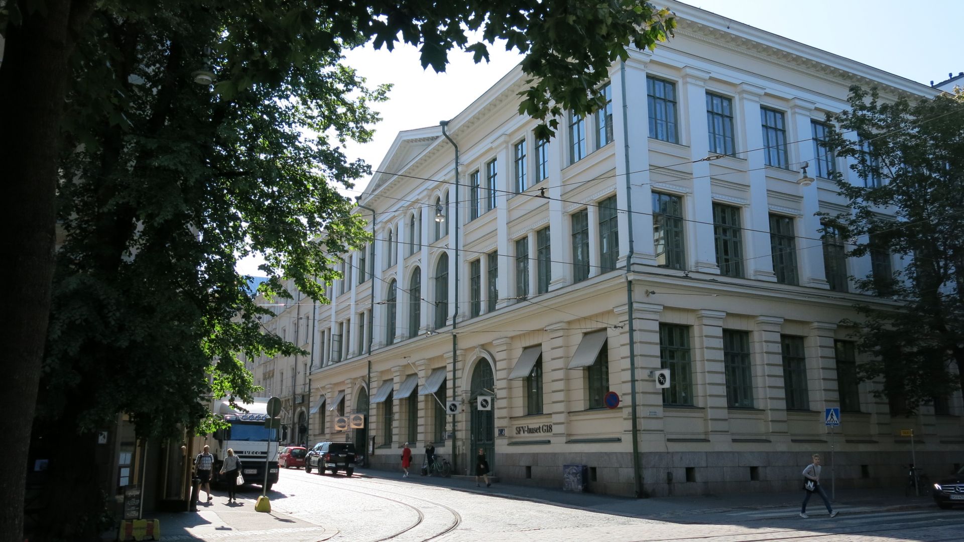 SFV-huset G18 fotograferat från andra sidan gatan en sommardag. I förgrunden syns gröna träd och gatan i korsningen Bulevarden - Georgsgatan. Huset är gult, med vitmålade pelare och grönt kring fönstren.