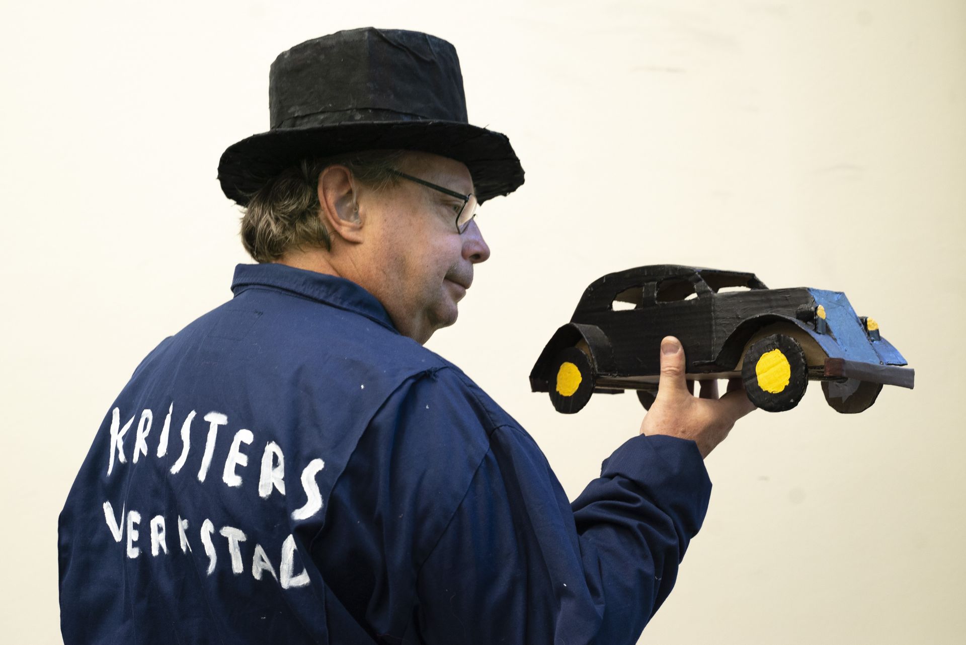 Skådespelare Krister Ekebom iklädd en svart hatt och en blå arbetshalare. Han står med ryggen snett mot kameran och håller en svart bil tillverkad av papp i handen. På hans rygg är texten Kristers verkstad målad. 