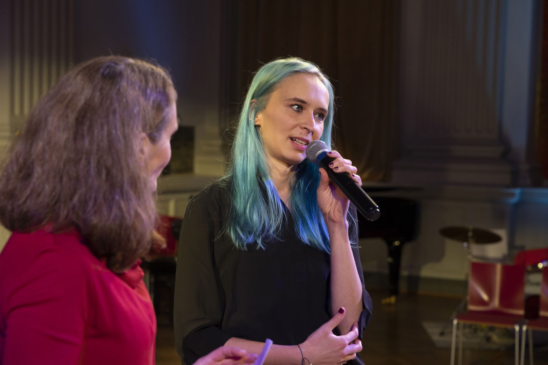 Emma Raunio sinisine hiuksineen kertoo esityksestä mikrofoni vasemmassa kädessään. Hänen oikealla puolellaan seisoo DuvTeaternin ohjaaja Mikaela Hasán.