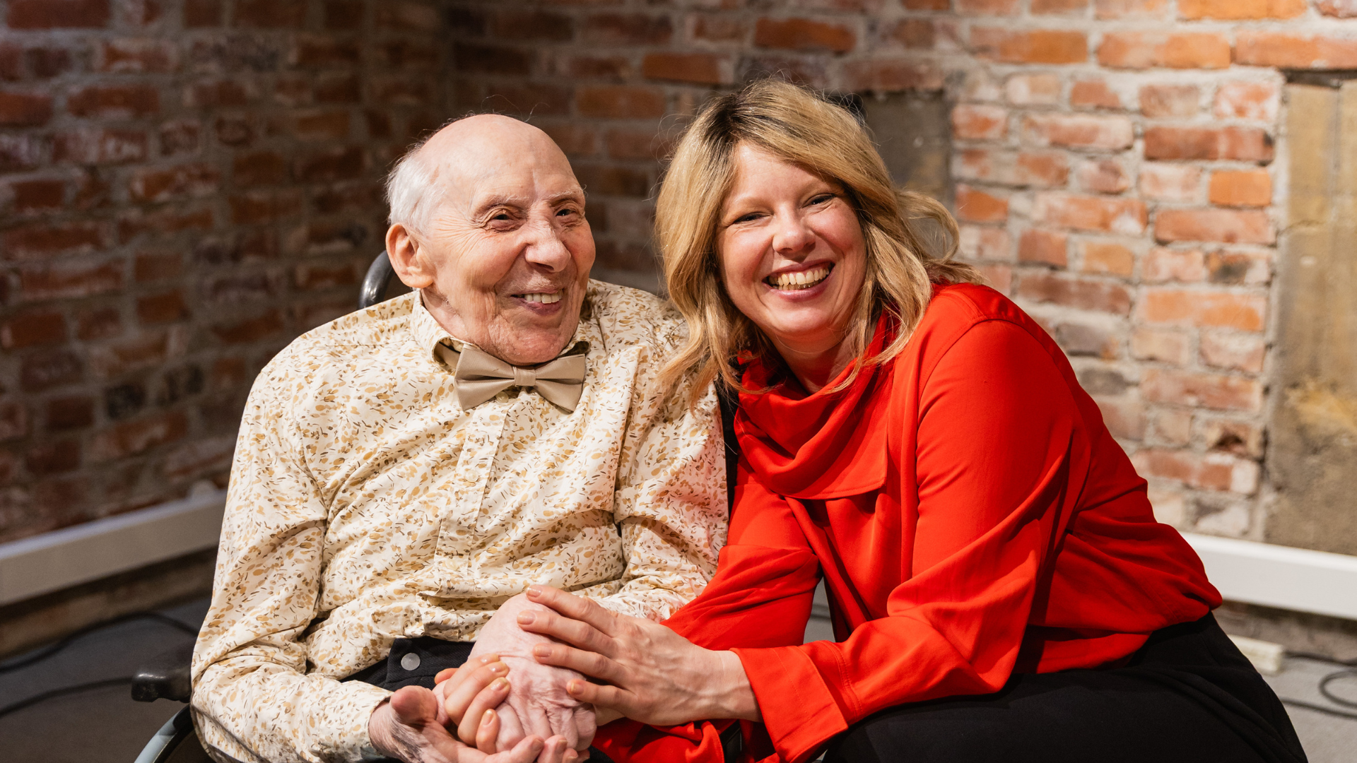 Två personer sitter framför en tegelvägg. En äldre man iklädd skjorta och fluga sitter bredvid en kvinna klädd i rött. De håller varandras händer och båda ler mot kameran.